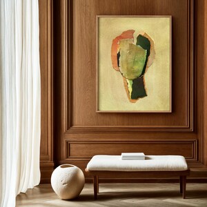 May include: Abstract art print in a wooden frame. The artwork features a collage of green, orange, and black shapes on a light yellow background. The print is displayed on a wood-paneled wall above a bench with a white cushion.