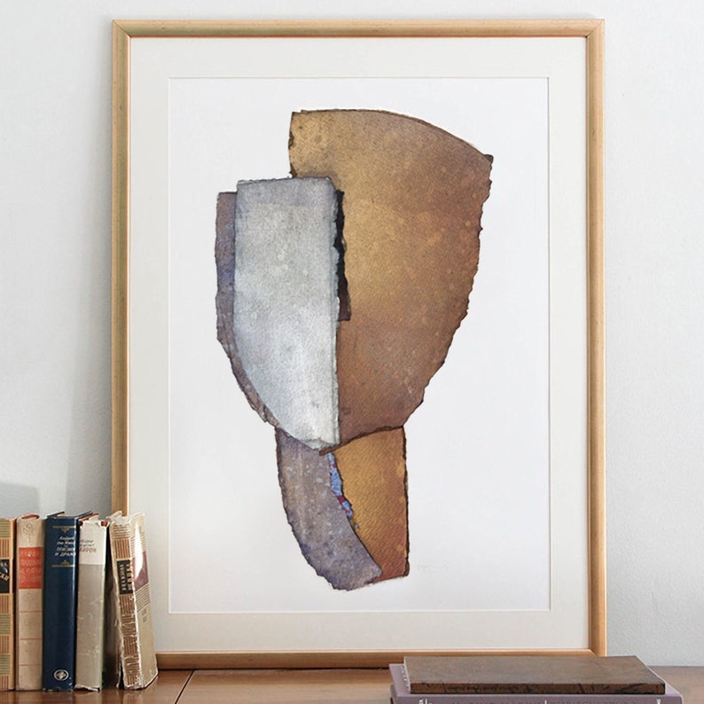 An interior design with old books and a gold framed high quality art print of an abstract portrait in beige to golden brown, dove gray and off-white, with black and sky blue accents. The torn paper layers have woven texture and rough, dark edges.