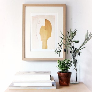 A wall art with a fine quality light wood framed art print of an abstract portrait. It is in a tender yellow pallet, with torn paper edges in red, a pencil drawn eye for an accent. Layers in desert sand cut out a head from a pale peach background.