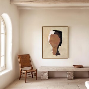 May include: An abstract painting of a face in a gold frame hangs on a cream-colored wall. The artwork features brown, black, and white shapes. A wooden chair with a woven back and a stone bench with a wooden bowl are in the room.