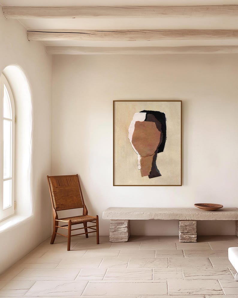 May include: An abstract painting of a face in a gold frame hangs on a cream-colored wall. The artwork features brown, black, and white shapes. A wooden chair with a woven back and a stone bench with a wooden bowl are in the room.