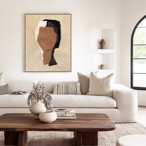 May include: A modern living room with a large abstract art piece in a gold frame. A white sofa is adorned with decorative pillows. A wooden coffee table displays books and vases. The room has a neutral color palette with natural light.