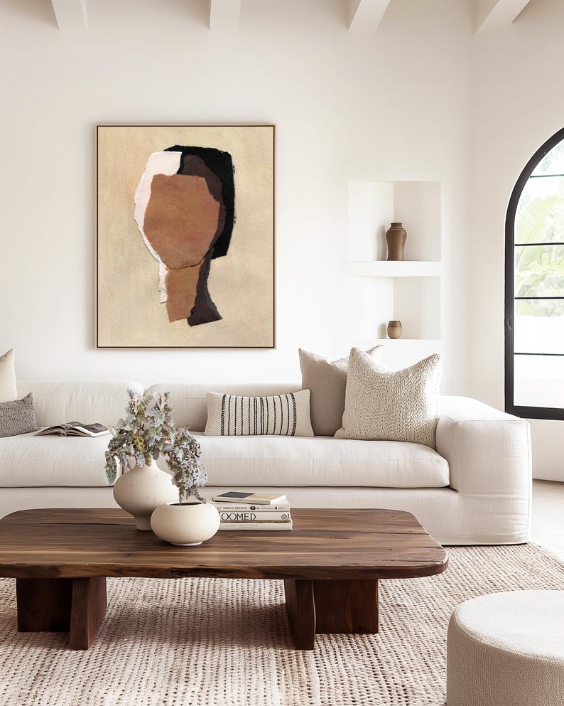 May include: A modern living room with a large abstract art piece in a gold frame. A white sofa is adorned with decorative pillows. A wooden coffee table displays books and vases. The room has a neutral color palette with natural light.