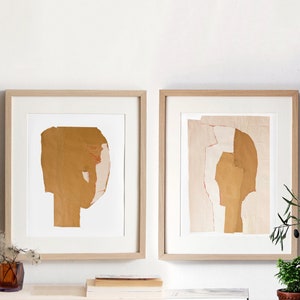 A neutral wall art with two maple wood framed high quality art prints of abstract portraits in pale peach and hues of desert sand yellow. Minimal eyes. The layers of ripped paper have rough edges. The set is balanced and neutral, best for wall decor.