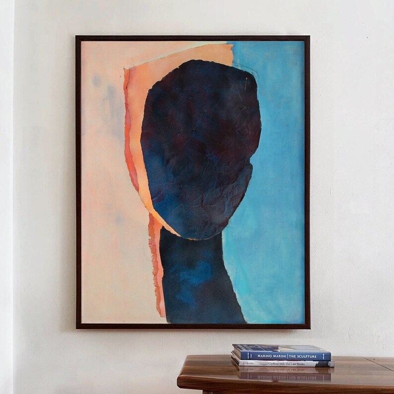 A wall decor with an art print of an abstract portrait in peach fuzz, shades of dark indigo blue on a pale salmon and sky blue background. The work is composed of three torn paper layers with rough edges and wrinkled texture in a black minimal frame.