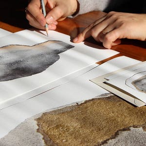 May include: A person is drawing on a white sheet of paper with a pencil. The paper has a brown and gray watercolor design on it. There are other sheets of paper with similar designs in the background.