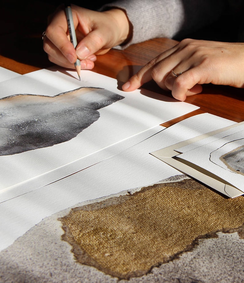 May include: A person is drawing on a white sheet of paper with a pencil. The paper has a brown and gray watercolor design on it. There are other sheets of paper with similar designs in the background.