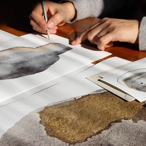 May include: A person is drawing on a white sheet of paper with a pencil. The paper has a gray and brown abstract design on it. There are other sheets of paper with similar designs on the table.