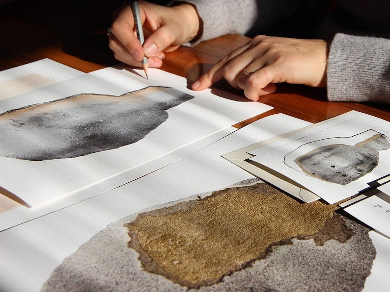 May include: A person is drawing on a white sheet of paper with a pencil. The paper has a gray and brown abstract design on it. There are other sheets of paper with similar designs on the table.