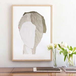 A decor with white flowers and  books on a lacquered wood shelve, and a light framed art print of an original abstract portrait in greige, of-white and ecru. The edges of the torn paper layers outline the head on white for a minimal graphic feeling.