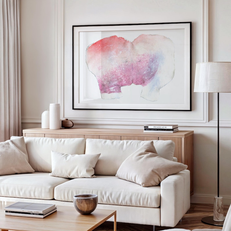 K&ouml;nnte beinhalten: Ein wei&szlig;es Sofa mit zwei Kissen, ein Couchtisch mit einer Schale und B&uuml;chern und ein gerahmtes abstraktes Aquarellbild an der Wand &uuml;ber einem Holzschrank mit B&uuml;chern.