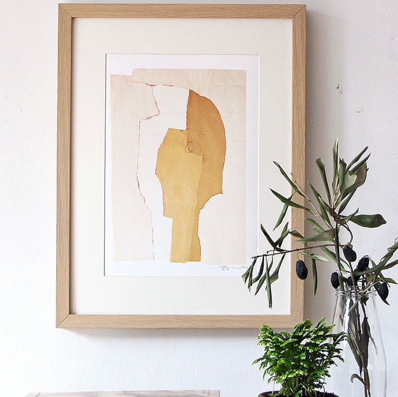 An accent decor with a high quality mat-framed art print of an abstract portrait. It has tender peach yellow pallet, torn paper edges in cherry red, a pencil drawn eye for an accent. Layers in desert sand cut out a head from a pale peach background.