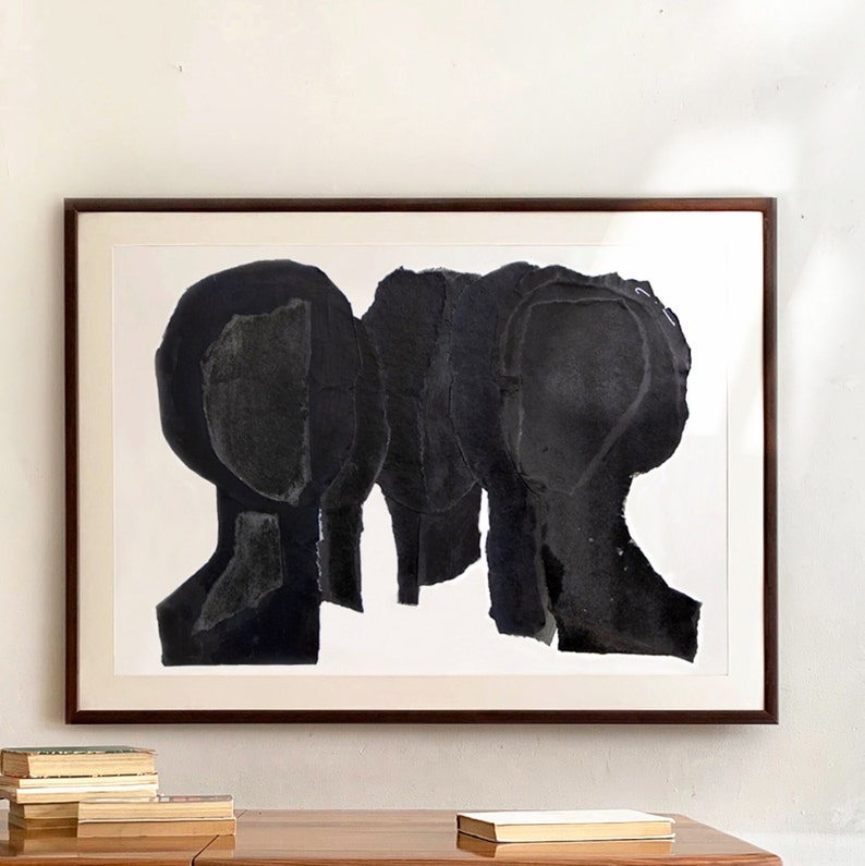 A wall decor with a large statement art in shades of brushed eerie black and grey and an oak table with some books. This dark wood framed group portrait has five abstract heads. The arch composed, overlapping layers are in opaque shades of black.