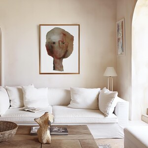 May include: A white couch with two white pillows in a living room with a framed abstract painting of a person's face and a lamp on the side.
