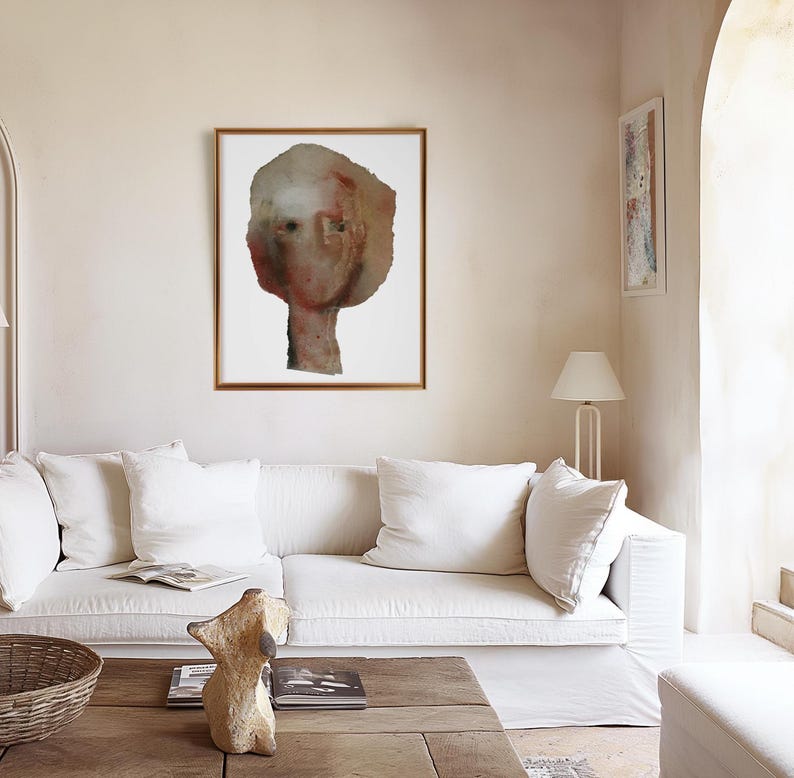 May include: A white couch with two white pillows in a living room with a framed abstract painting of a person's face and a lamp on the side.