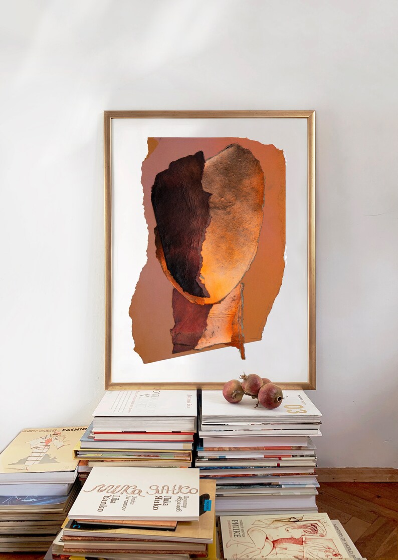 A studio decor with a high quality gold framed large wall art print of an abstract portrait in shades of vivid brown, varying from dark wood to blazing orange. A reddish drop spills out from the torn paper, random-shaped, faded persimmon background.