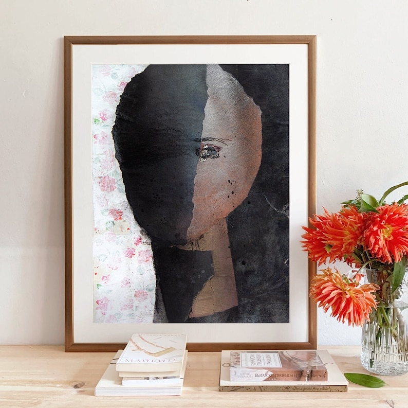 May include: A framed abstract painting of a face with a black and brown color scheme. The face is partially obscured by a black background. The painting is set against a white wall with a vase of orange flowers in the foreground.