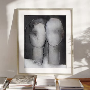A wall decor with a light framed art print of a noir abstract portrait of a grayscale love couple on spray-painted black background and white passe-partout. The heads have torn paper layers with exposed grungy edges and two roughly outlined eyes.