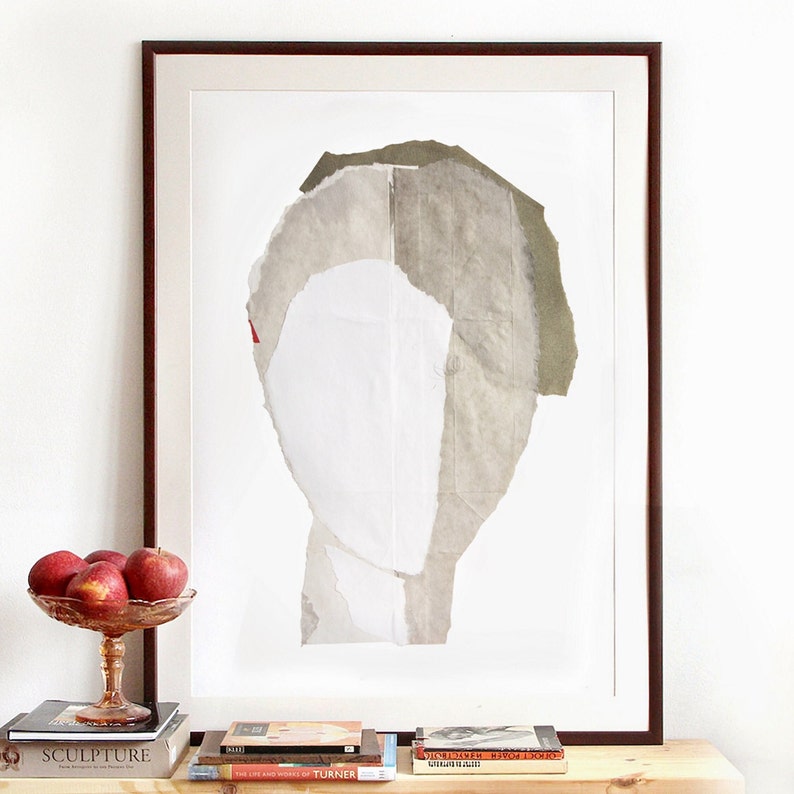 A decor of a dark framed art print of an original abstract portrait in greige, of-white, icy white and ecru on a pile of art books. The edges of the thick paper torn layers outline the head on white for a minimal graphic feeling with the beige paper.