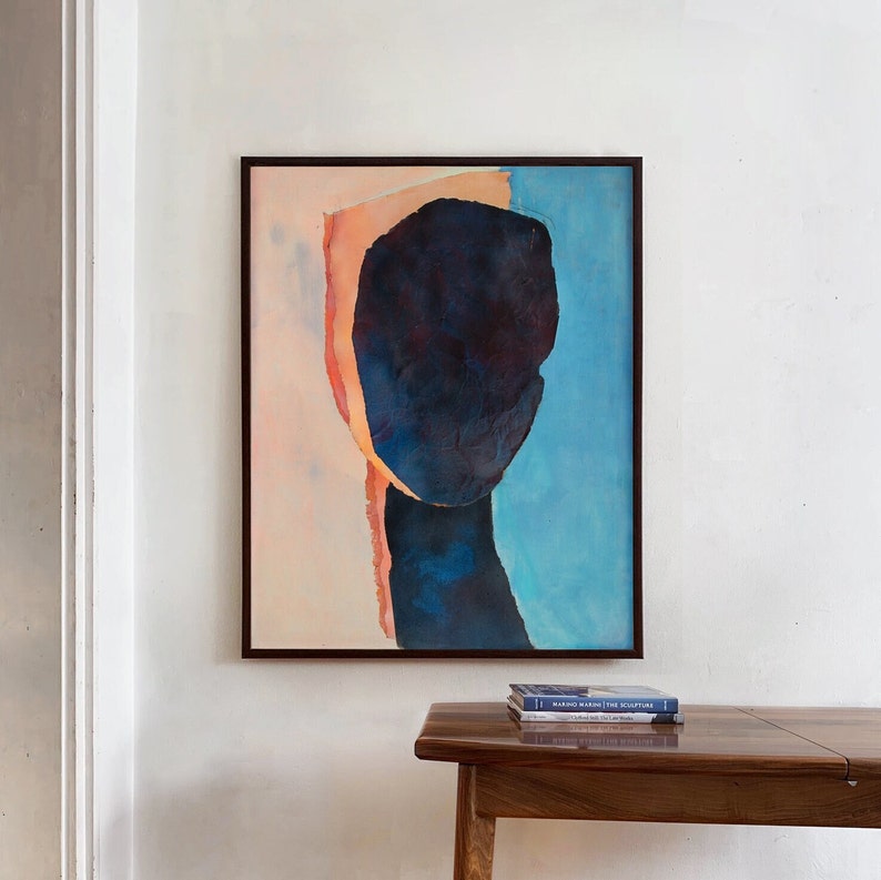 A wall decor with an art print of an abstract portrait in peach fuzz, shades of dark indigo blue on a pale salmon and sky blue background. The work is composed of three torn paper layers with rough edges and wrinkled texture in a black minimal frame.