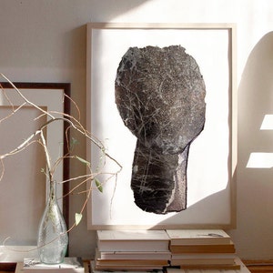 A visual set up with a modern light wood framed art print of an abstract head in shades of graphite grey, with silver spray painted accents. The torn paper layers have rocky texture, webbed with light grey and sprinkled in white and charcoaled edges.