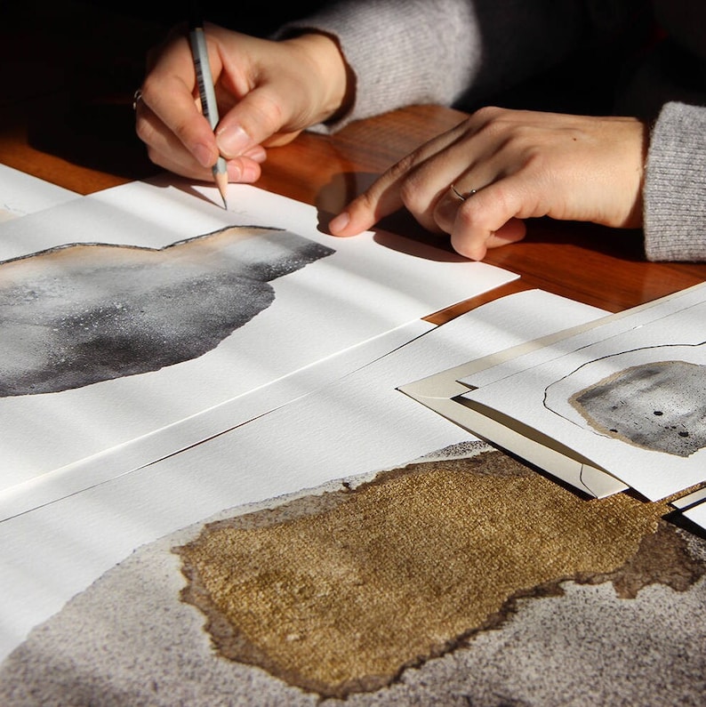 Puede incluir: Se muestran las manos de una persona dibujando en una hoja de papel blanca con un l&aacute;piz. El papel est&aacute; sobre una mesa de madera. Hay otras hojas de papel con dibujos en el fondo.