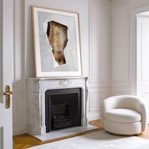May include: A white marble fireplace with a black fireplace insert and a framed abstract art print above it. The fireplace is in a room with white walls and a white armchair.