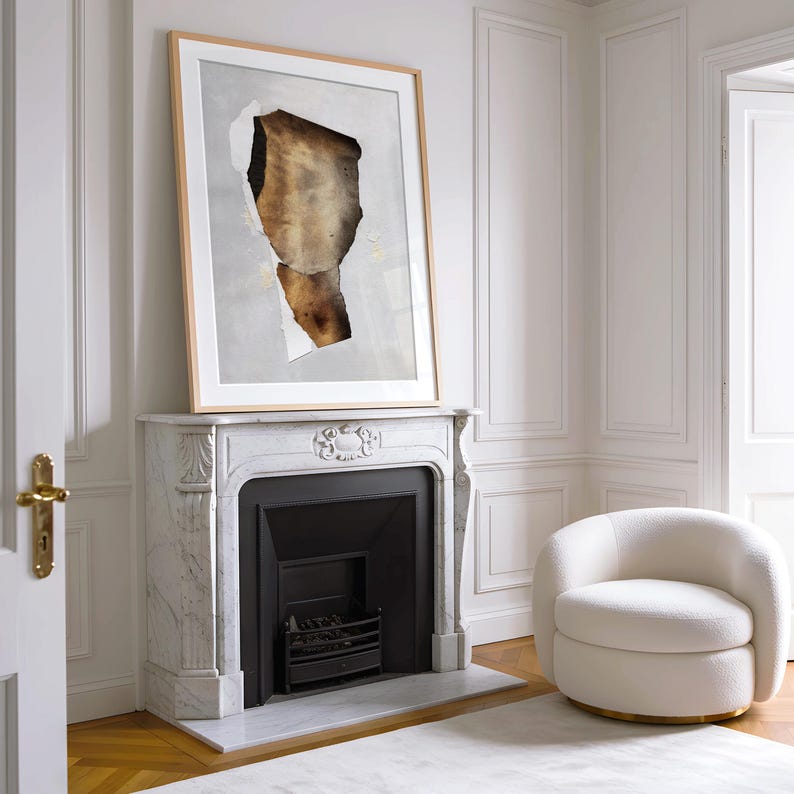 May include: A white marble fireplace with a black fireplace insert and a framed abstract art print above it. The fireplace is in a room with white walls and a white armchair.