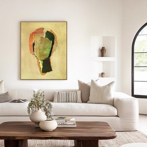 May include: Abstract art print in a gold frame. The artwork features a collage-like design with green, orange, and dark green shapes on a beige background. The painting hangs above a white sofa with decorative pillows and a wooden coffee table.