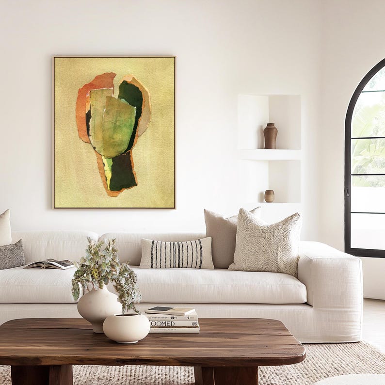 May include: Abstract art print in a gold frame. The artwork features a collage-like design with green, orange, and dark green shapes on a beige background. The painting hangs above a white sofa with decorative pillows and a wooden coffee table.