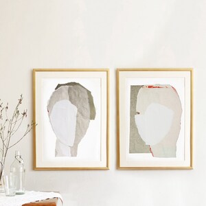 A neutral wall art with two gold framed high quality art prints of abstract portraits in pale white, pink and greige. One has a red minimal eye. The layers of ripped paper has coarse contours. The set is balanced and neutral, best for wall decor.