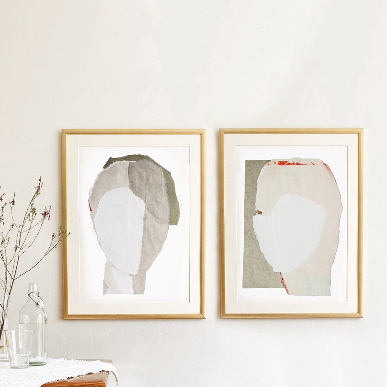 A neutral wall art with two gold framed high quality art prints of abstract portraits in pale white, pink and greige. One has a red minimal eye. The layers of ripped paper has coarse contours. The set is balanced and neutral, best for wall decor.