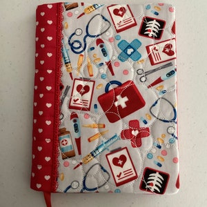 May include: A red and white quilted notebook cover with a medical theme. The cover features a variety of medical symbols, including a stethoscope, a first aid kit, a thermometer, and a syringe. The cover is trimmed with a red and white ribbon.