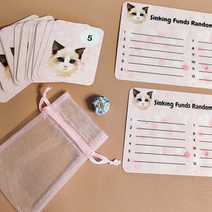 以下が含まれることがあります： 猫のイラストと「Sinking Funds Randomizer」というテキストが書かれたピンクのカード12枚セット。カードはピンクと白のサイコロとピンクのオーガンザバッグを背景に積み重ねられています。