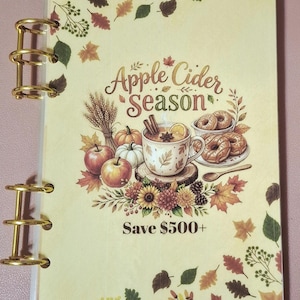 May include: A clear plastic binder with gold-tone metal rings featuring a cream-colored cover with autumn-themed illustrations of apples, pumpkins, donuts, and flowers. The text reads Apple Cider Season and Save $500+. The design includes fall leaves.