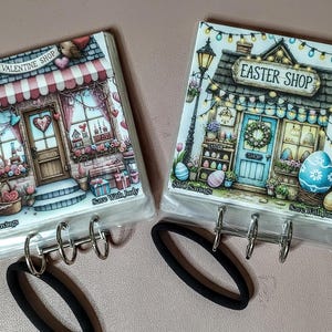 May include: Two decorative binder covers, one labeled "Valentine Shop" and the other "Easter Shop." Each features a detailed illustration of a shop with holiday-themed decorations, including hearts and Easter eggs. The covers are attached to binder rings.