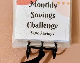 May include: A white and pink monthly savings challenge binder with gold hearts and the text "Monthly Savings Challenge $300 Savings".