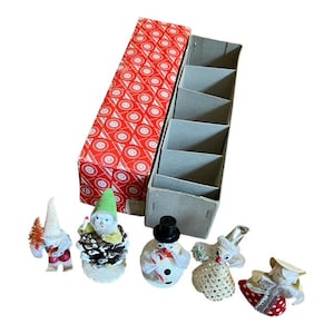 Peut inclure: Ensemble de décorations de Noël vintage, comprenant des bonhommes de neige et des gnomes, présentées à côté de leur boîte d'origine. La boîte est rouge avec un motif circulaire blanc. Les ornements sont petits et colorés, avec des accents de paillettes.