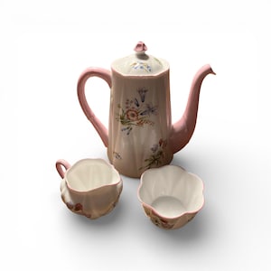 May include: A ceramic tea set in pink and white. The set comprises a teapot with a lid, a creamer, and a sugar bowl. The teapot and other items are decorated with floral patterns.