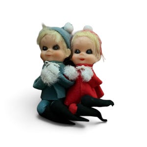 May include: Two small elf figurines with blonde hair and painted facial features. One elf wears a blue felt outfit with a matching cap and white pom-poms, while the other wears a red felt outfit with a matching cap and white pom-poms. Both have black pointed shoes.