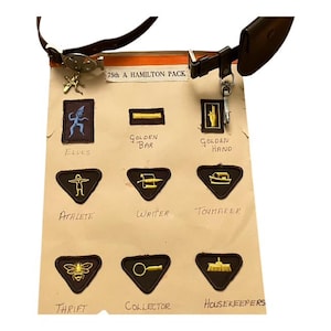 May include: A display of vintage patches featuring gold and black designs, such as an elf, a golden bar, a hand, and other symbols. The text "75th A HAMILTON PACK" is present. The patches are attached to a brown leather strap.