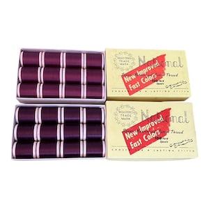 Peut inclure: Deux boîtes de bobines de fil bordeaux avec des bandes blanches, à côté de deux boîtes de fil de marque "National". Les boîtes présentent le texte "New Improved Fast Color Cotton Thread" et "100 Yard Spools".