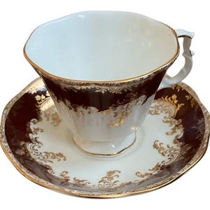 May include: A vintage teacup and saucer set. The teacup has a white interior and a dark brown exterior with gold accents. The saucer is white with a dark brown border and gold floral designs. The set has a gold trim.