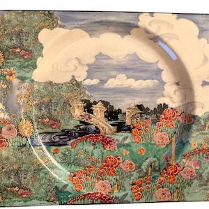May include: A square ceramic plate with a raised rim features a colourful landscape scene. The scene depicts a bridge over a river, surrounded by trees and flowers in shades of pink, red, green, and blue. The sky is a light blue with white clouds.