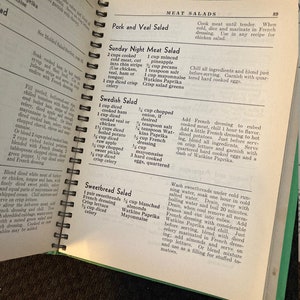May include: Open vintage cookbook with a green cover and a recipe for Swedish Salad. The recipe includes ingredients like cooked ham, veal, chicken, potato, apple, onion, and mayonnaise.