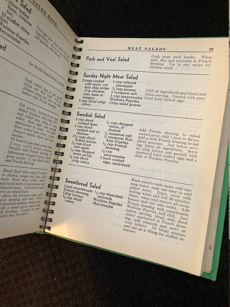 May include: Open vintage cookbook with a green cover and a recipe for Swedish Salad. The recipe includes ingredients like cooked ham, veal, chicken, potato, apple, onion, and mayonnaise.