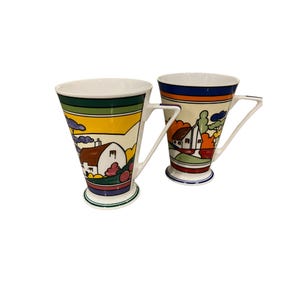 May include: Two white ceramic mugs with colourful geometric designs. Each mug features a stylised landscape with a house, trees, and a sky. The mugs have a unique angled handle.