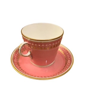 May include: A pink teacup and saucer set with gold detailing. The teacup has a curved handle and a white interior. The saucer is round and complements the teacup's design. The set is likely made of porcelain.