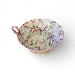 May include: A vintage teacup with a pink handle and rim, decorated with a floral pattern. The cup showcases pink and white flowers with green foliage on a pale blue backdrop. A classic teacup design.