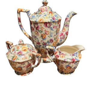 May include: A vintage floral tea set featuring a teapot, sugar bowl, and creamer. The set has a cream background with a colorful floral pattern in shades of pink, yellow, and blue. The teapot has a tall, curved spout and a matching lid.